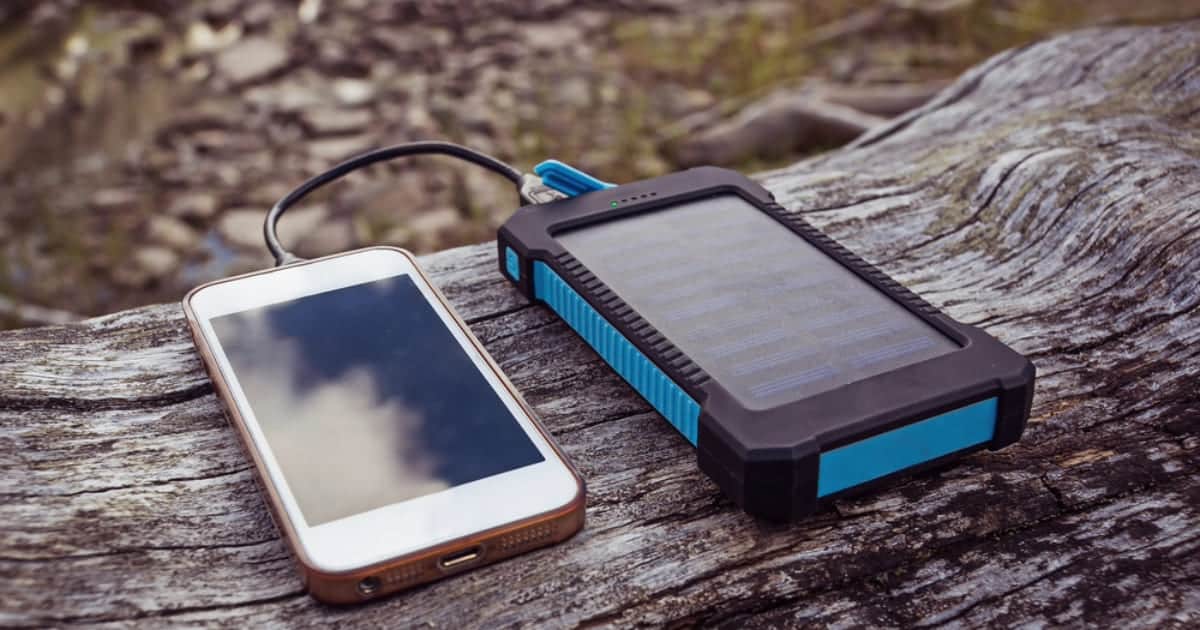 Power bank solaire X-Moove connecté à un smartphone posé sur bois flotté en pleine nature avec lac de montagne
