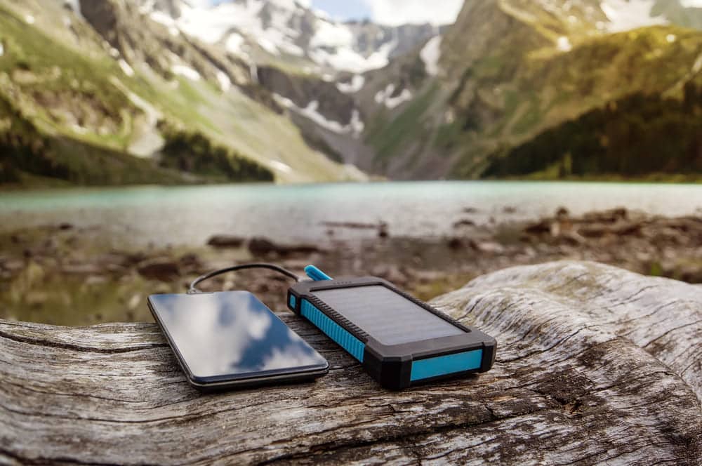 Batterie externe solaire X-Moove bleue rechargeant un smartphone lors d'une randonnée en montagne au bord d'un lac