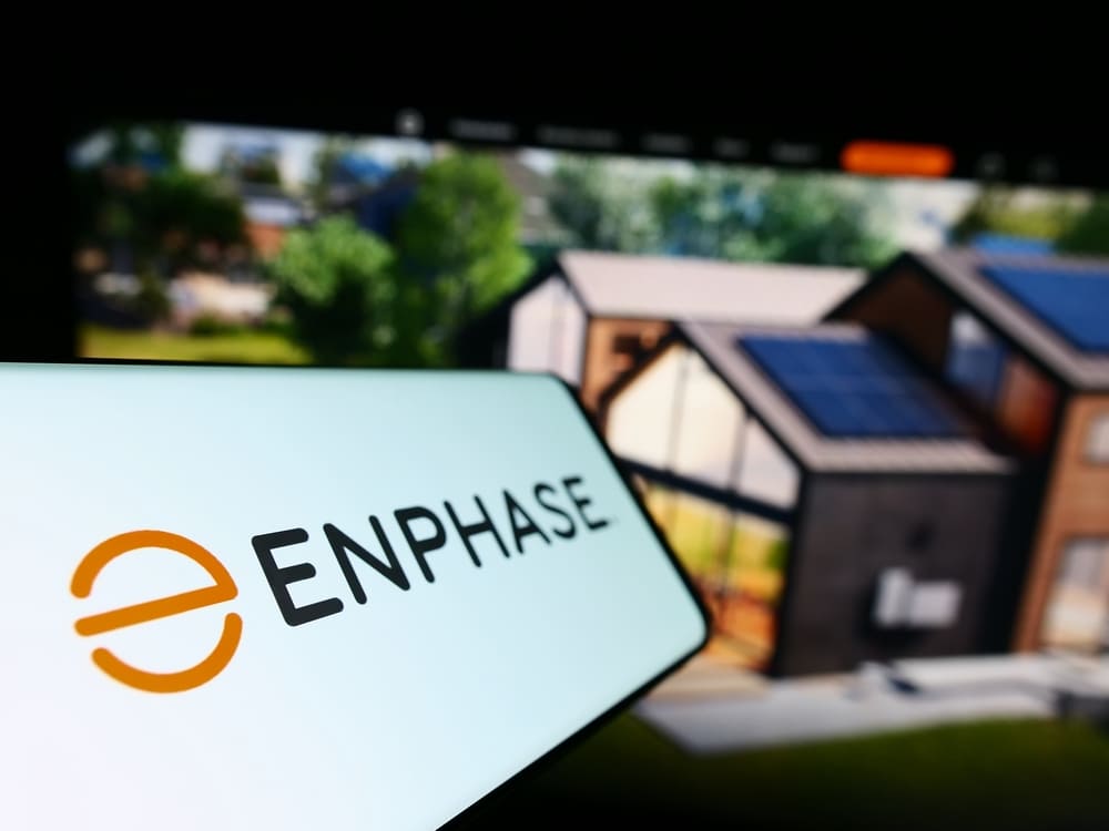 Logo Enphase sur smartphone devant une maison équipée de panneaux solaires, leader des micro-onduleurs photovoltaïques